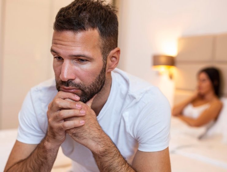 A man with erectile dysfunction sitting on the bed with his wife in the background.