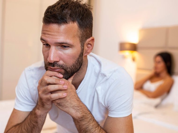 A man with erectile dysfunction sitting on the bed with his wife in the background.
