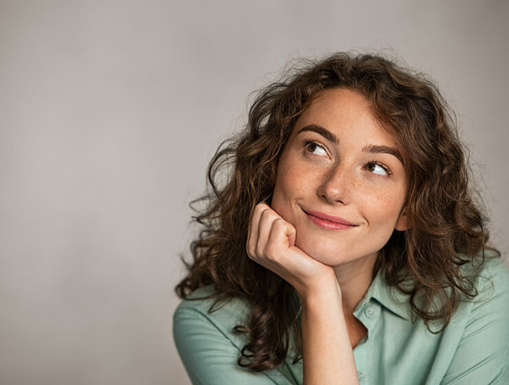 Young woman with curly hair looks thoughtfully, facing the side with her chin in her hand.
