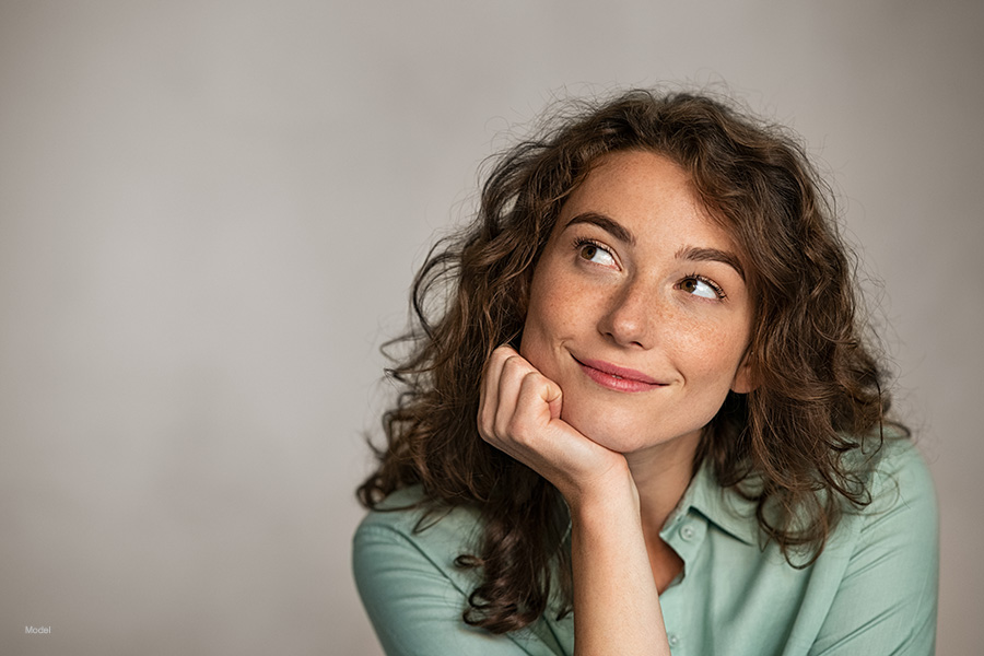 Young woman with curly hair looks thoughtfully, facing the side with her chin in her hand.