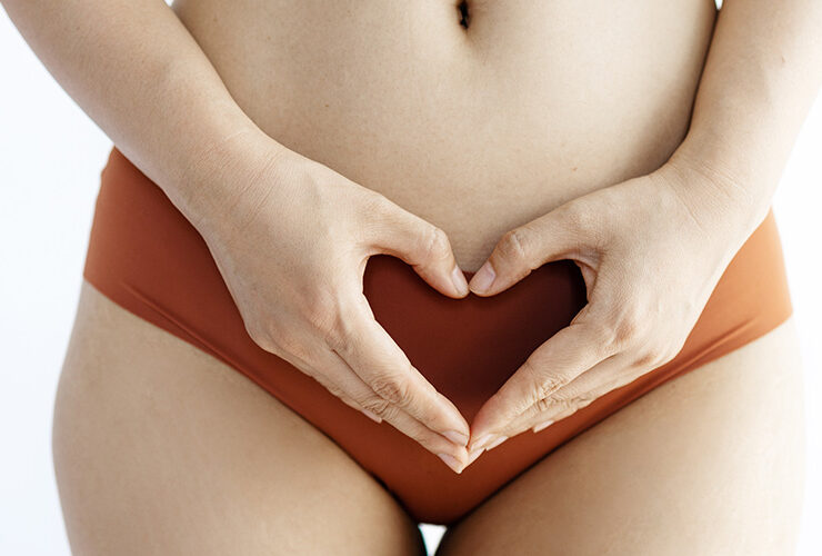 A woman wearing orange underwear and holding her hands in a heart shape across her pelvis.