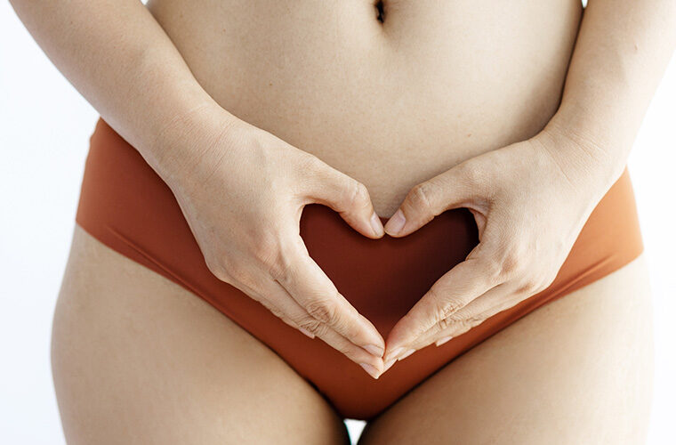 A woman wearing orange underwear and holding her hands in a heart shape across her pelvis.